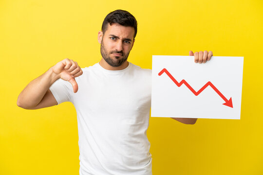 Young handsome caucasian man isolated on yellow background holding a sign with a decreasing statistics arrow symbol and doing bad signal