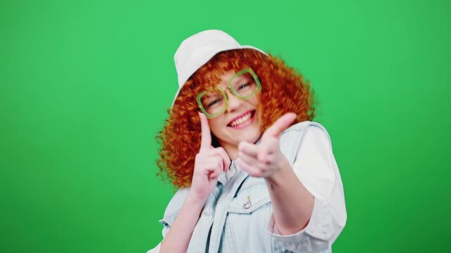 Young playful redhead curly woman in eyeglasses and panama hat dancing and gesturing gotcha, green chroma key background