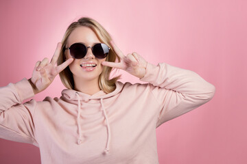 Naklejka premium beautiful young woman with blonde hair wearing dark sunglasses makes a peace gesture, two fingers up, with two hands. In a pink hoodie on a pink background, studio portrait