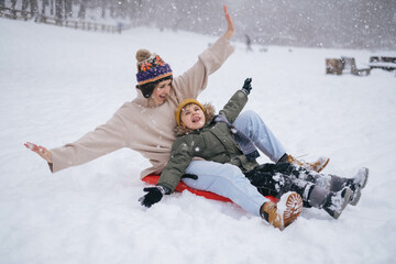 Fototapeta na wymiar Mother and son having fun in winer snowy park, riding on sled, family travel on vacation, christmas. Winter clothing, winter sports. Lifestyle, kids, parenting, winter nature.