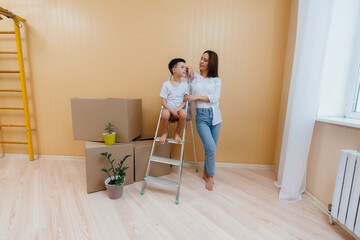 A young woman and her son are standing in front of the boxes and enjoying the housewarming after moving in. Housewarming, delivery and freight transportation, purchase of real estate.