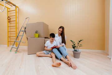 Fototapeta na wymiar A young woman with her son is sitting in front of the boxes and rejoicing at the housewarming after moving in. Housewarming, delivery and freight transportation, purchase of real estate.