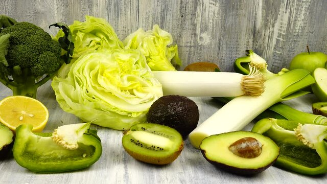 Vegetables and fruits of green color whole and halves lie on a wooden table.  Avocado and lemon, leek, broccoli, kiwi, apples and green bell pepper iceberg lettuce are on a table.