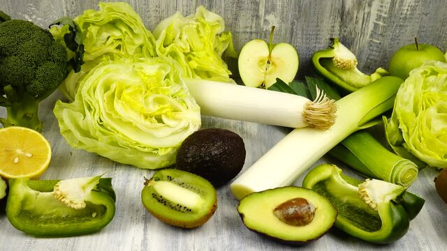 Vegetables and fruits of green color whole and halves lie on a wooden table.  Avocado and lemon, leek, broccoli, kiwi, apples and green bell pepper iceberg lettuce are on a table.
