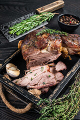 Roasted mutton lamb leg sliced in a wooden tray with meat cleaver. Black wooden background. Top view