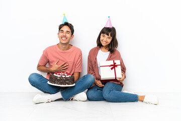 Young mixed race couple celebrating a birthday sitting on the floor isolated on white background keeping the arms crossed while smiling
