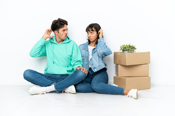 Young couple making a move while picking up a box full of things sitting on the floor isolated on white background having doubts while scratching head