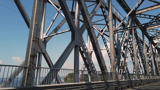 metal bridge. rough iron constructions against blue sky and river, filmed in motion from a car. side view. Railway and road bridge across the river. Silhouette of crossing steel beams.