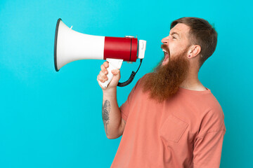 Young reddish caucasian man isolated on blue background shouting through a megaphone to announce something in lateral position