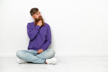 Young caucasian reddish man sitting on the floor isolated on white background having doubts and with confuse face expression