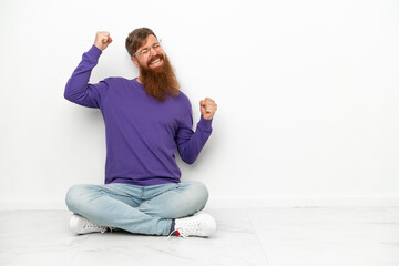 Young caucasian reddish man sitting on the floor isolated on white background celebrating a victory