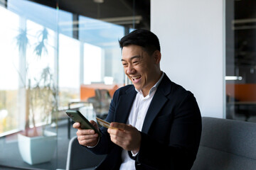 Happy asian man shopping online, businessman in office, shopping in online store using credit card, happy with the loan