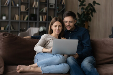 Relaxed happy bonding young family couple using computer applications, planning travel vacation booking tickets, shopping in internet store, watching movie or web surfing, sitting on sofa at home.