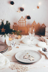 Organization of a festive Christmas table. Plate with wooden decorations, cutlery, champagne glass and cardboard house. Tablescapes on white wooden table. Vertical view