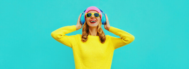 Portrait of happy smiling young woman with headphones listening to music wearing colorful yellow sweater on blue background
