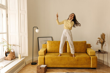 Full-length european young woman is jumping on couch and listening to music through headphones in living room. Slim, dark-haired girl has fun on her weekends. Lifestyle concept