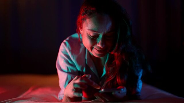 adult woman is surfing social media in internet by smartphone in night, lying in bed in darkness