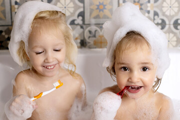 Two little girl brushing teeth seating in bathroom with bath foam. Kids personal care. Family fun