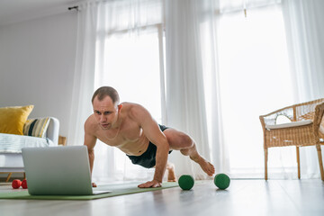 Home workout in the living room. Young muscular body man making spiderman pushups following online a fitness trainer using a laptop. Healthy lifestyle and modern online technology concept. © Soloviova Liudmyla