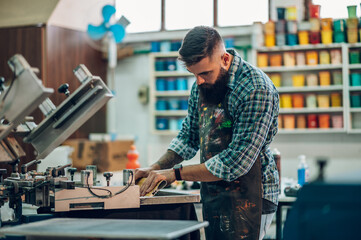 Male worker pressing ink on frame while using the printing machine in a workshop