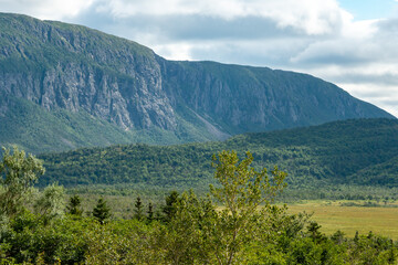 Gros Morne mountain on the Northern Peninsula of Newfoundland is flat on top with a hiking trail up. The sky is cloudy with blue spots. There are hills, lush marsh, and trees in front of the mountain 