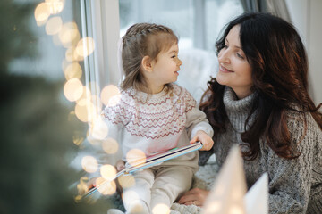 Young grandmother play and read book with her adorable grandaugher nea fir tree. Christmas mood