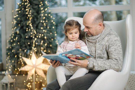 Young grandfather play and read book with her adorable grandaugher nea fir tree. Christmas mood