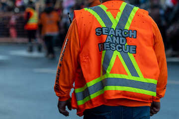 A male adult stands among a crowd wearing a bright orange vest with the words search and rescue. There's a grey and yellow x on the back of the industrial jacket. There are people in the background.