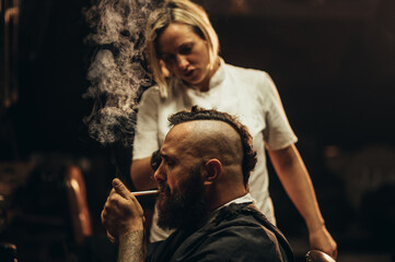Young bearded man with a mohawk getting haircut by hairdresser and smoking a cigarette