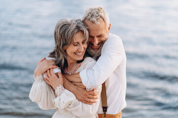 Romantic senior couple is embracing on seashore. Man hugs woman from behind.