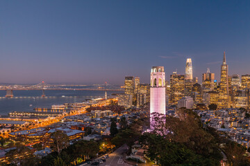 Nighttime aerial view of the San Francisco skyline with Coit Tower prominent in the frame. Bay Bridge in the background.