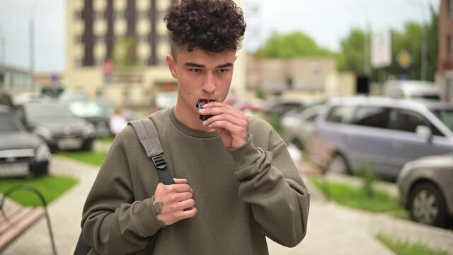 A young guy smokes an electronic cigarette, releasing steam. Slow motion, stylish guy walk around town
