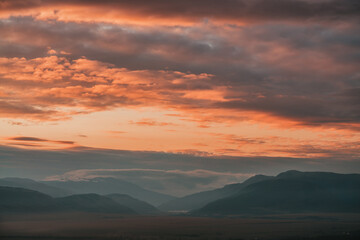 Obraz premium Scenic dawn mountain landscape with orange low clouds in valley among mountains silhouettes under cloudy sky. Vivid sunset or sunrise scenery with low clouds in mountain valley in illuminating color.