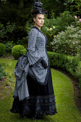 A Victorian woman wearing a striped silk polonaise and black skirt and standing in a beautiful...