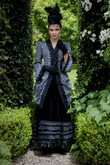 A Victorian woman wearing a striped silk polonaise and black skirt and standing in a beautiful...