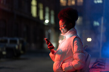 Happy african american woman standing on street at night, holding smartphone, calling taxi or...
