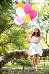 Cheerful beauty woman holding balloons relax sitting under big tree in green park with happiness. Woman Hands holding vibrant air balloons play on birthday party happy time summer on sunshine outdoor