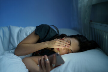 Tired woman lying in bed sleepless holding smartphone in hand reading about sleep disorder and...