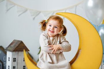 A beautiful two-year-old girl is sitting on a wooden decorative yellow moon in the studio. Portrait of a happy child on holiday.