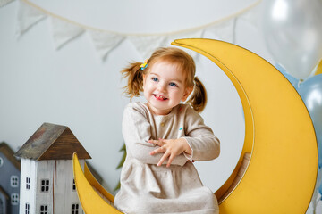 A beautiful two-year-old girl is sitting on a wooden decorative yellow moon in the studio. Portrait...