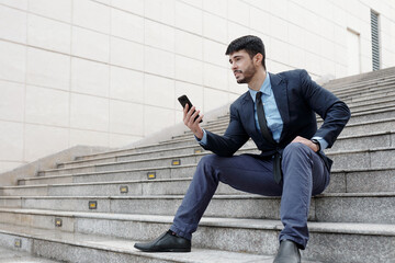 Confident young entrepreneur sitting on steps outdoors and reading article on smartphone online