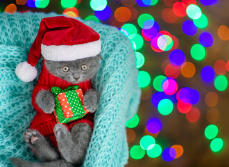 Surprised funny tiny kitten wearing warm sweater  and red santa hat lies inside basket and hugs  gift box. Top down view. Empty space for text
