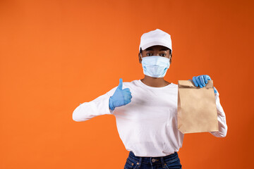 African-American delivery girl in white cap, rubber gloves and protective mask from viruses holding a package and showing her thumbs up on orange background with copy space