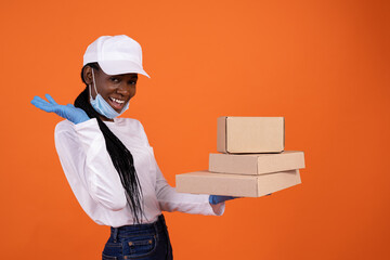 delivery, pandemic, helthcare and people concept - a happy African-American female courier in a pulled down protection mask and gloves holding boxes in the palm on orange background with copy space