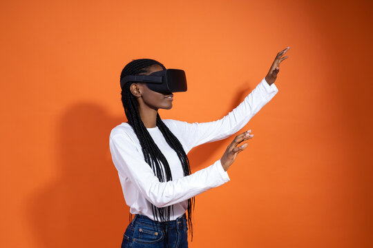 A young African-American woman wearing vr glasses sees something and tries to touch it with her hands on orange background with copy space