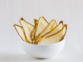 Dried thin slices, green pear fruit chips in white ceramic bowl. Healthy food and snacks. Fruits are bright orange in color. Side view, close up, Light background. Minimalism