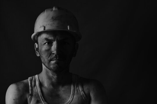 Dirty coal miner in a yellow hard hat on a dark background in a black and white photo.