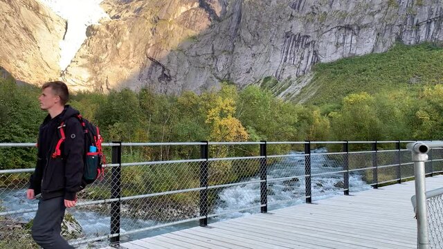 A young man walks relaxed on a bridge over a mighty river with the Briksdalsbre glacier in the background