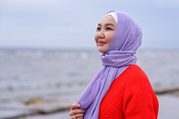 Portrait of beautiful happy pretty Asian Muslim young woman, Islamic girl is wearing on head hijab scarf, smiling, walking on the beach breathing deep fresh air. Traditional Muslim Arabian clothes.