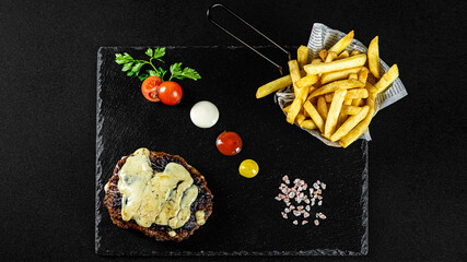 Grilled Gurmanska Pleskavica  with cheese, fries in a basket on a black plate with sauces and fresh veggies
